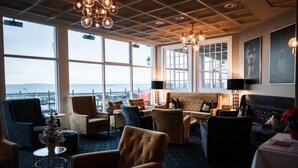 A cozy lounge area with comfortable seating, large windows offering a view of the sea, and a modern chandelier. - Grand Hotel Åsgårdstrand - Unike Hoteller (Horten)