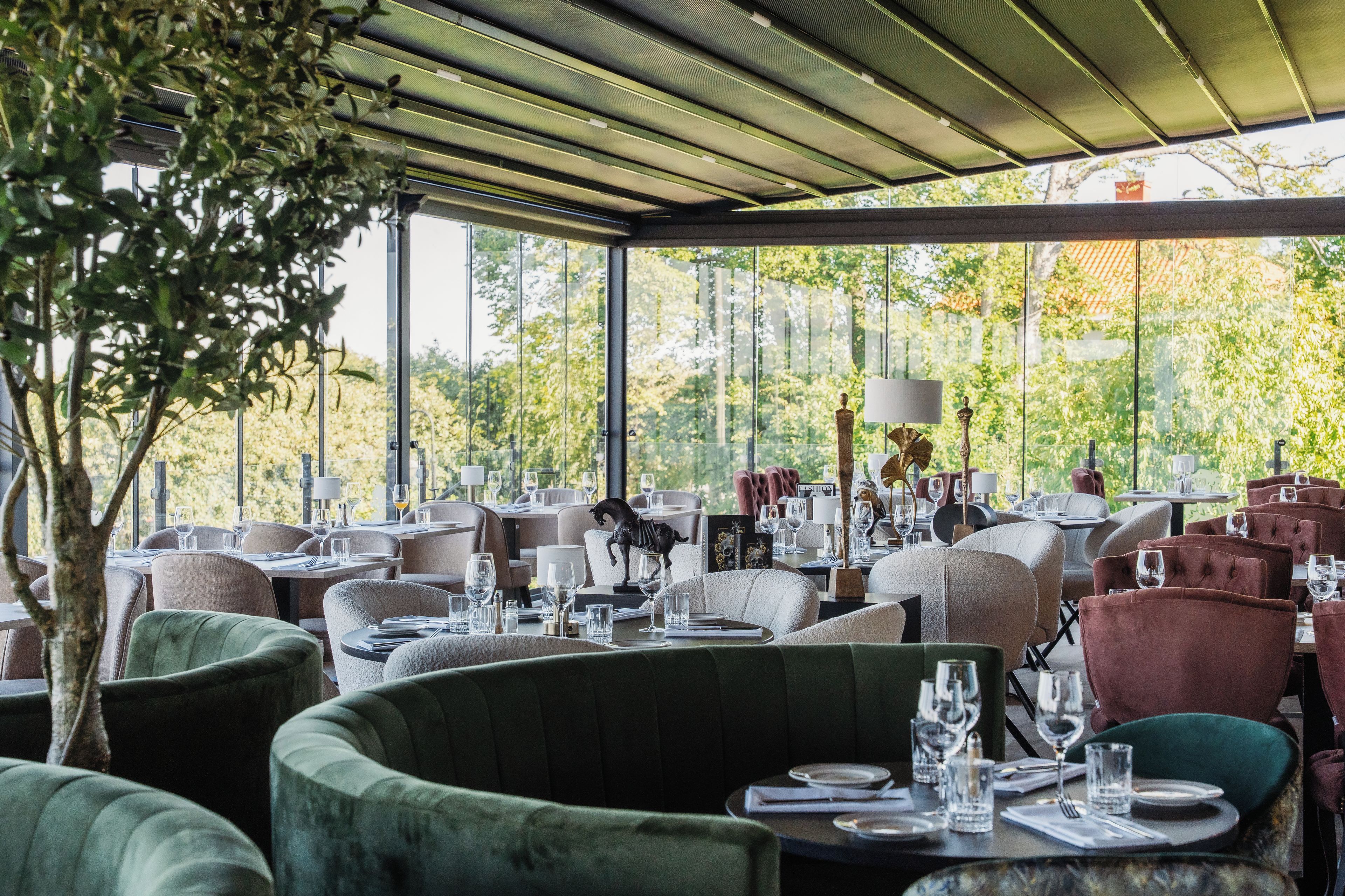 An indoor dining area with tables set for service, large windows offering a view of trees, and a ceiling with a grid-like pattern.