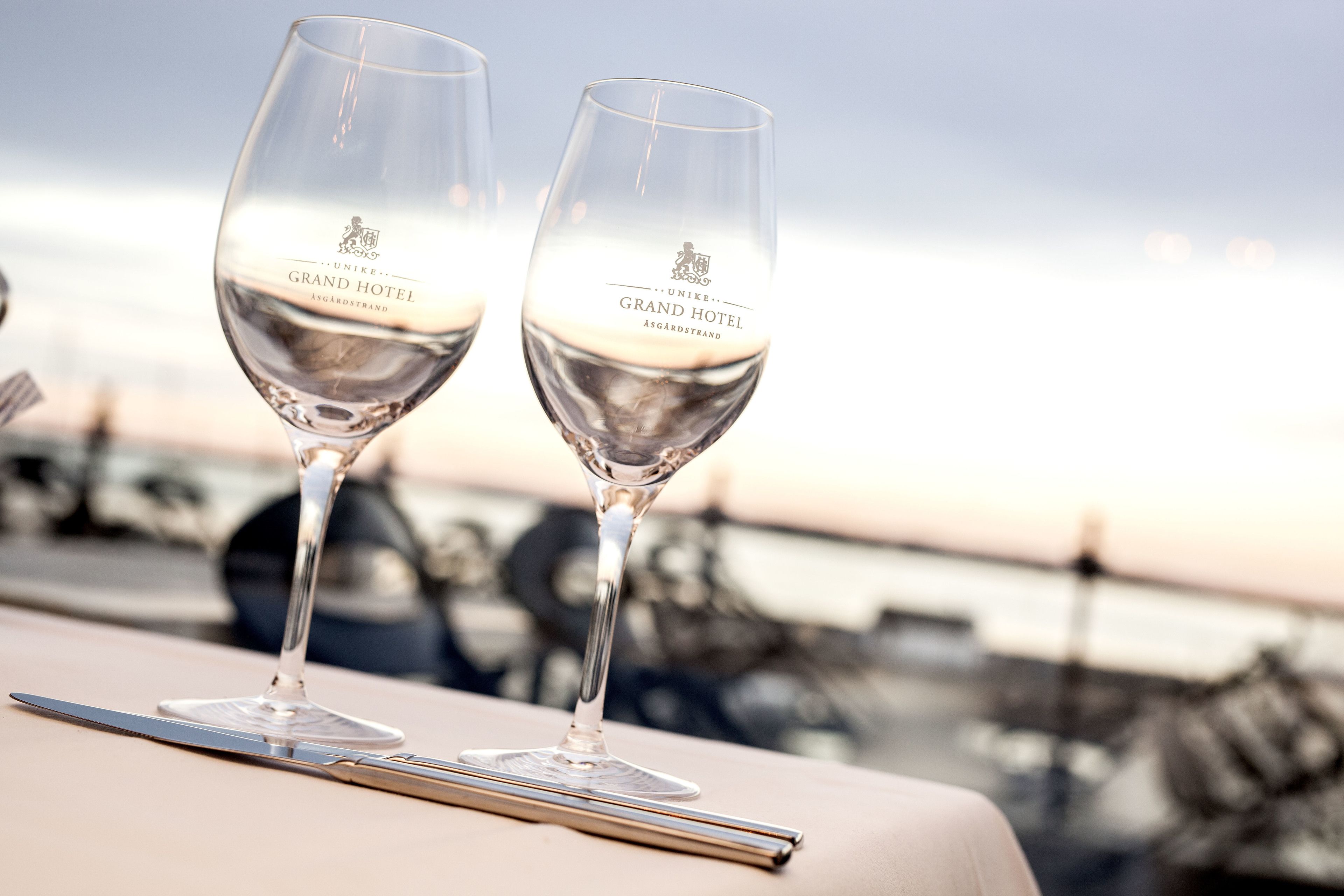 Dos copas de vino con la inscripción "UNIKe. GRAND HOTEL ASGARDSTRAND" sobre una mesa, con vista al mar y embarcaciones al fondo.