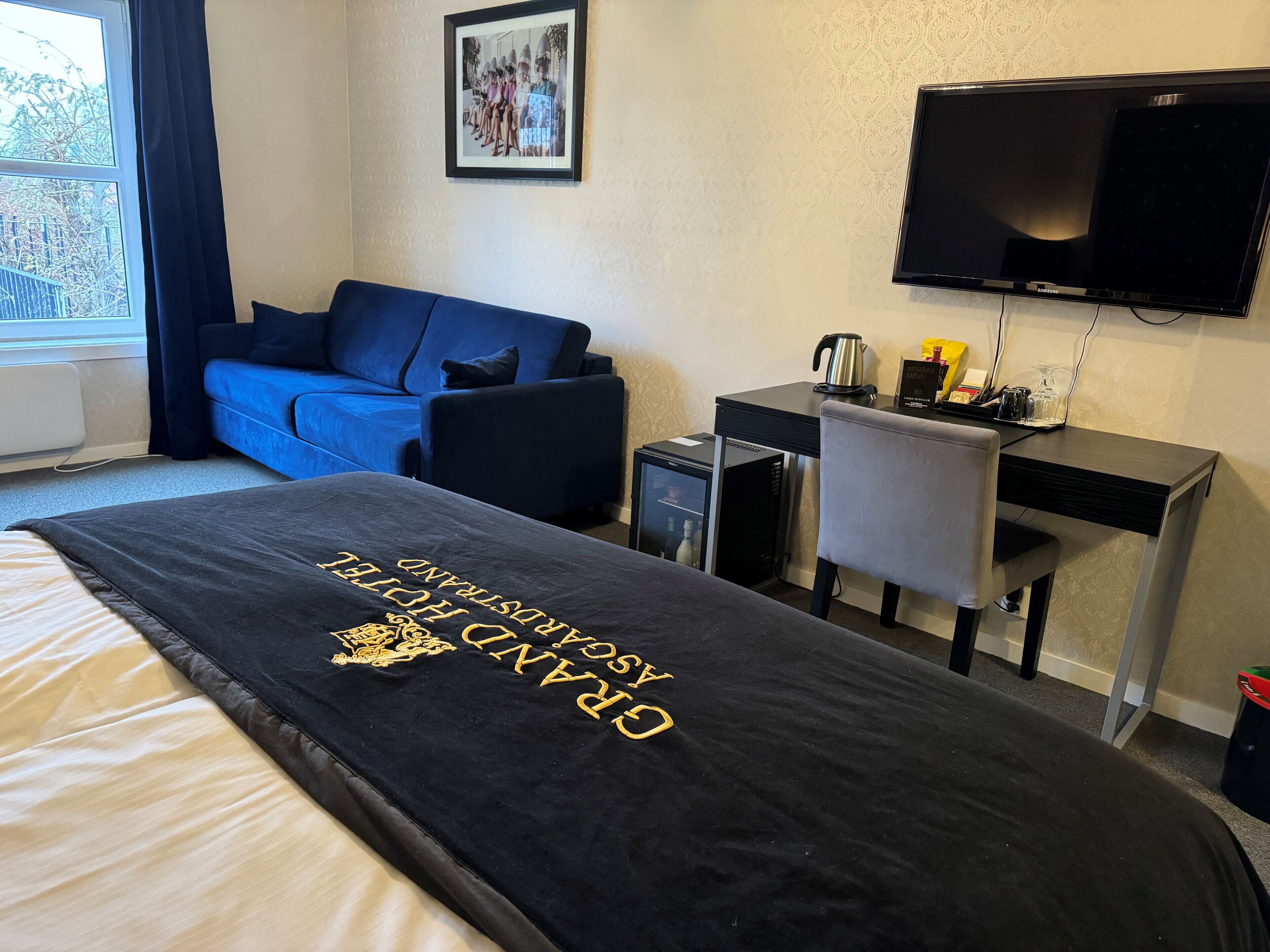 Habitación de hotel con cama, sofá azul, escritorio con computadora, televisor y ventana con cortinas.