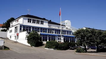 Un edificio de dos pisos con fachada blanca, ventanas grandes y una bandera roja en la parte superior.