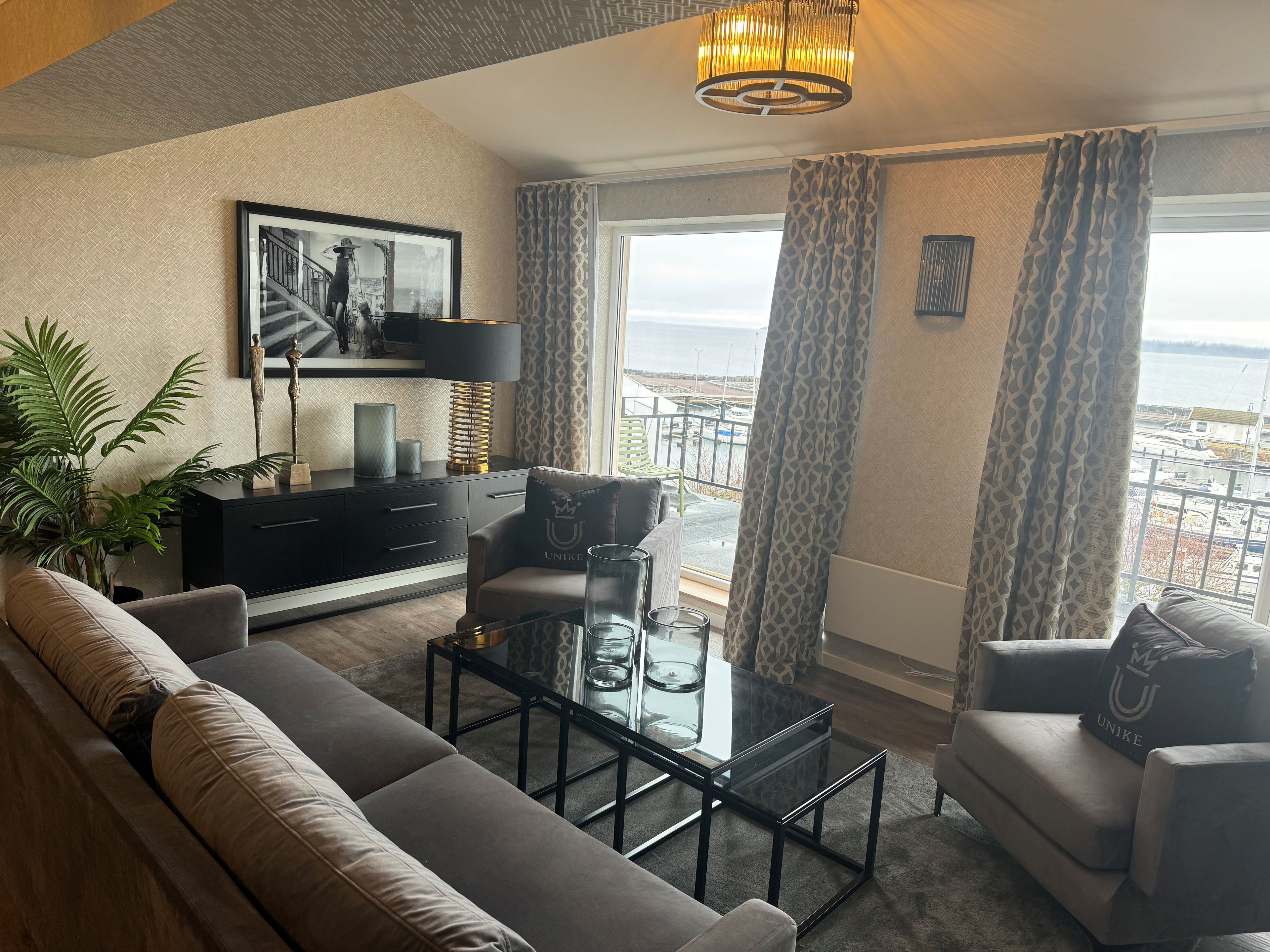 A modern living room with a sofa, armchairs, a glass coffee table, a black side table, a lamp, and a framed picture on the wall.