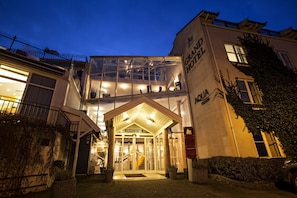 Entrada de un hotel con fachada de vidrio, una escalera que conduce a la recepción y un cartel con el nombre 'Grand Hotel'.