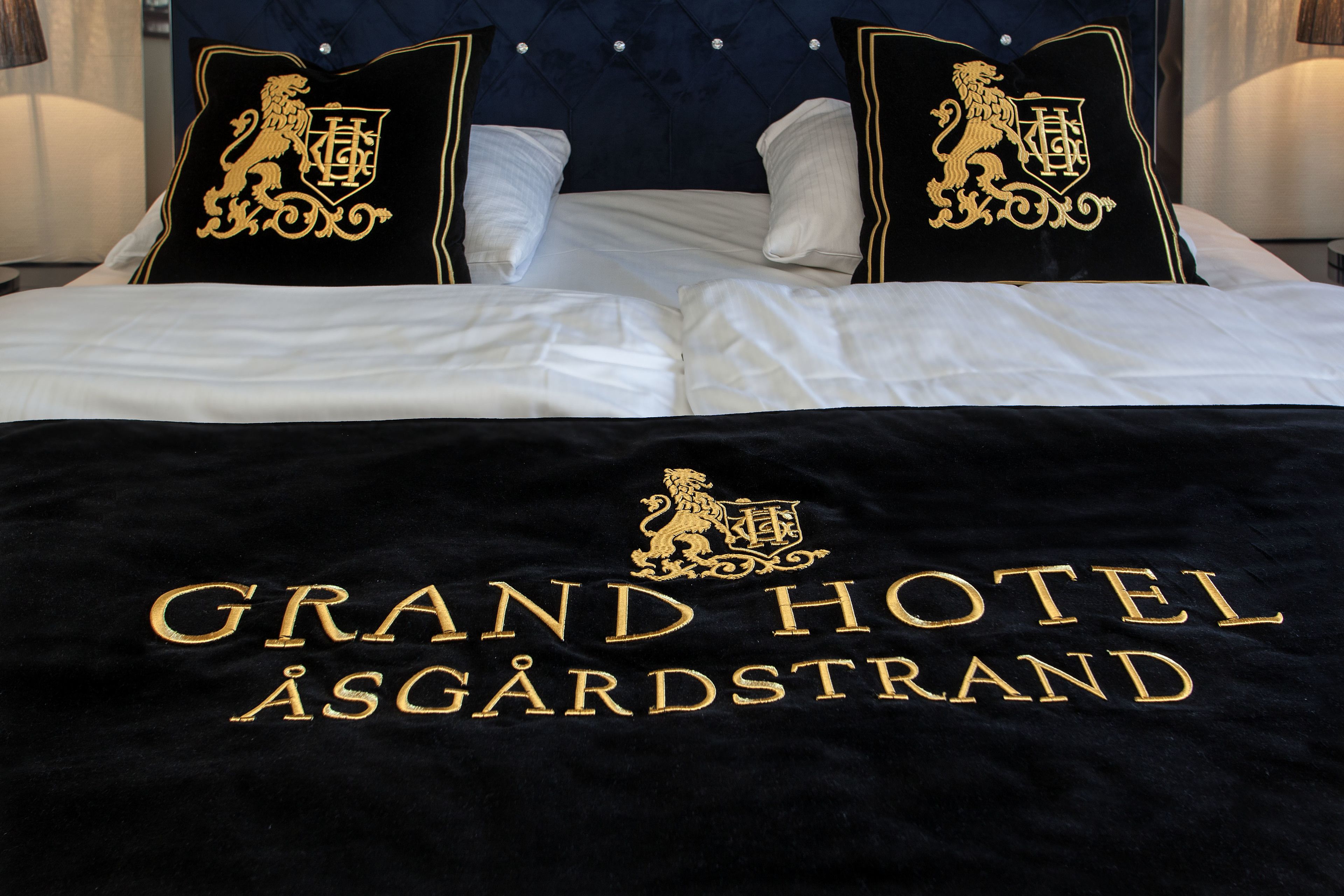 En seng med sort sengetæppe, der viser Grand Hotel Åsgårdstrands logo og navn.