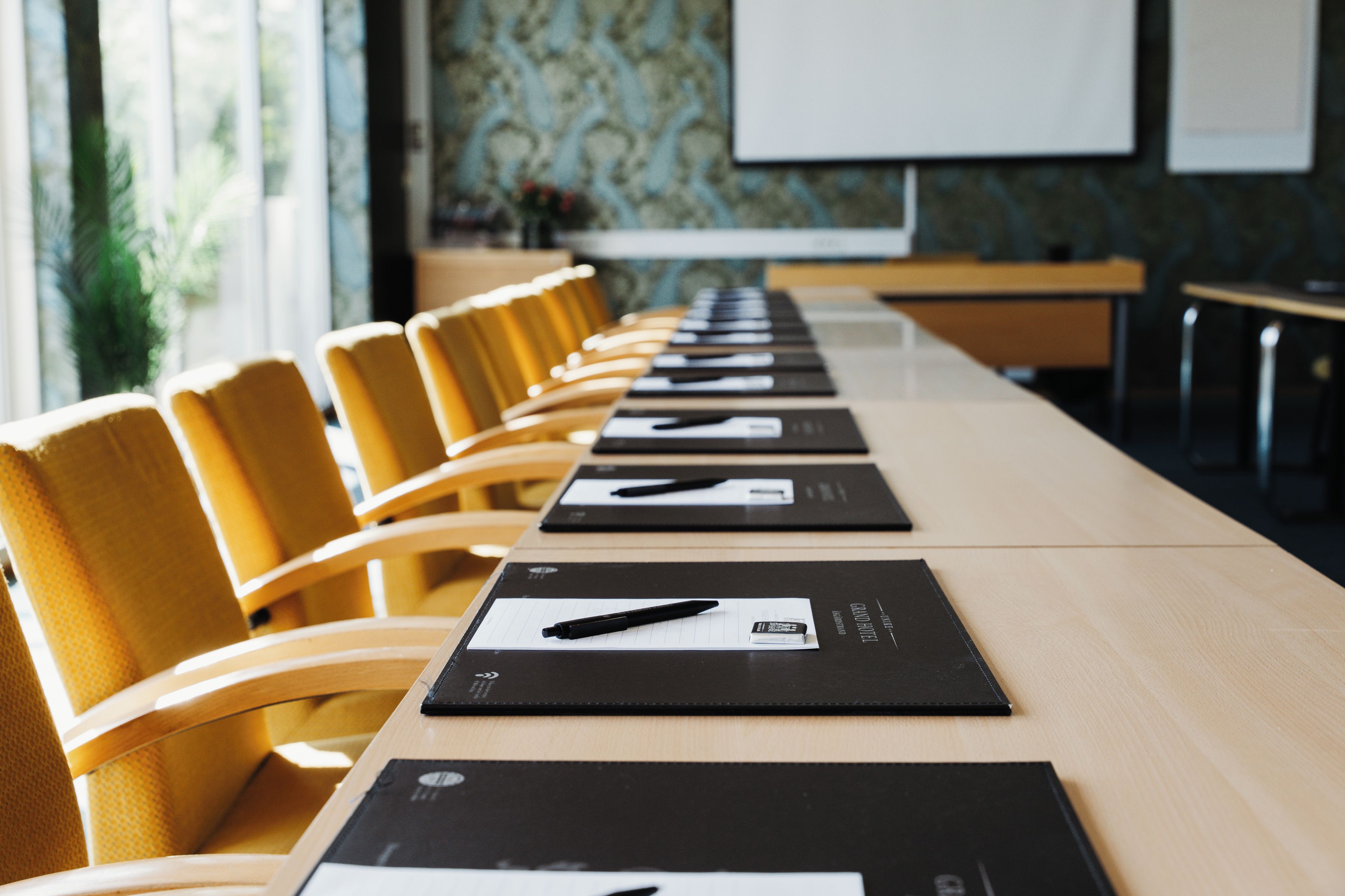 Een vergaderruimte met een lange tafel, gele stoelen en zwarte naamplaatjes met pennen.
