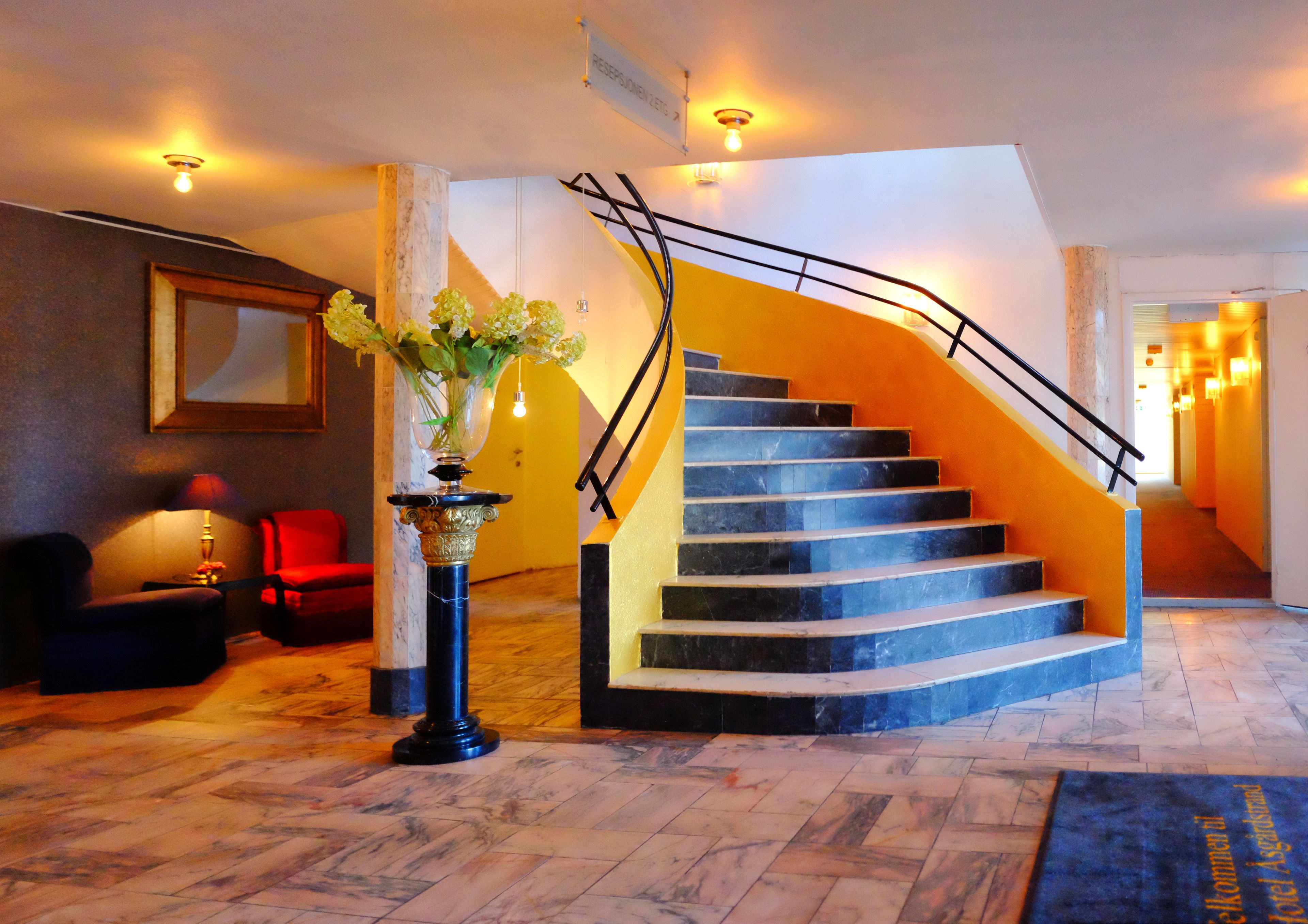 Un vestíbulo de hotel con suelo de mármol, una escalera con barandillas negras, una silla roja y un jarrón con flores.