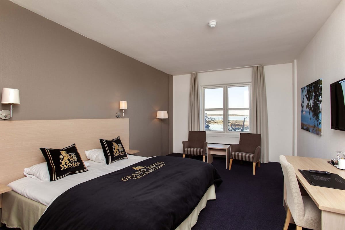 A hotel room with a bed, two chairs, a desk, a window with a view, and a mounted TV.