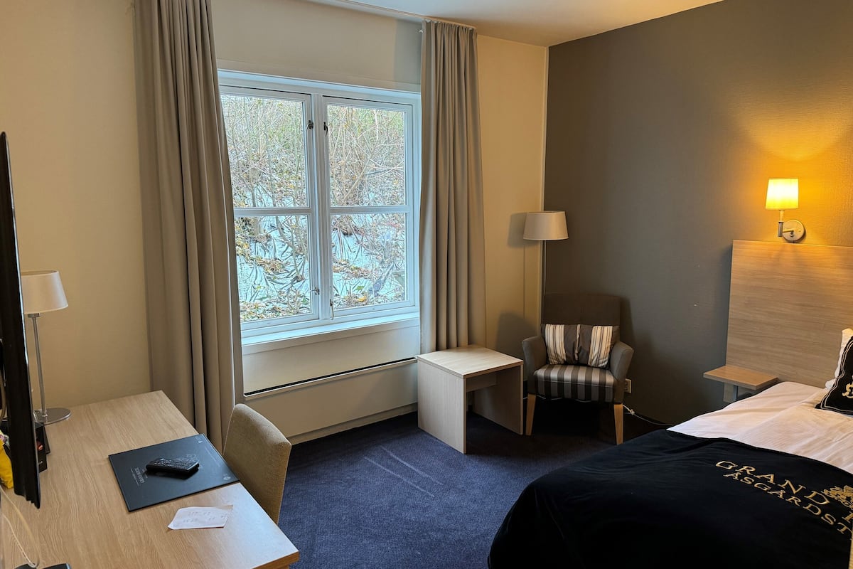 A hotel room with a bed, a desk, a chair, a window with curtains, and a lamp.