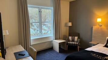 A hotel room with a bed, a desk, a chair, a window with curtains, and a lamp.