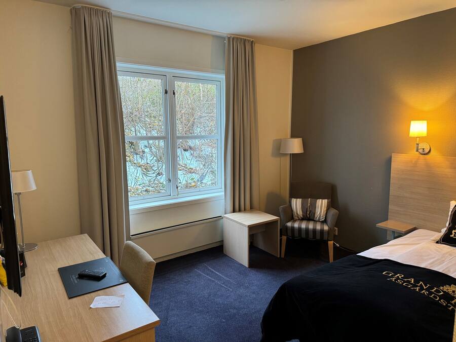 A hotel room with a bed, a desk, a chair, a window with curtains, and a lamp.