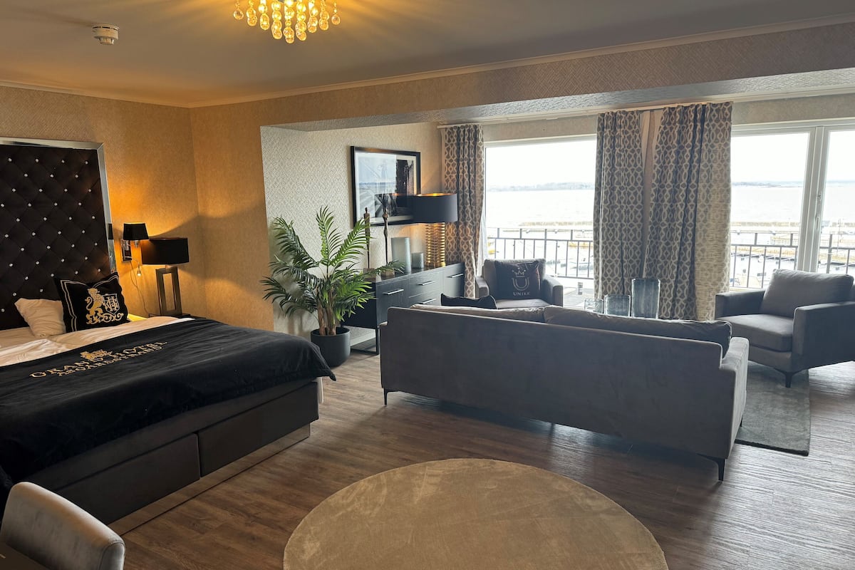 A modern hotel room with a bed, two sofas, a dining table, and a large window with a view.