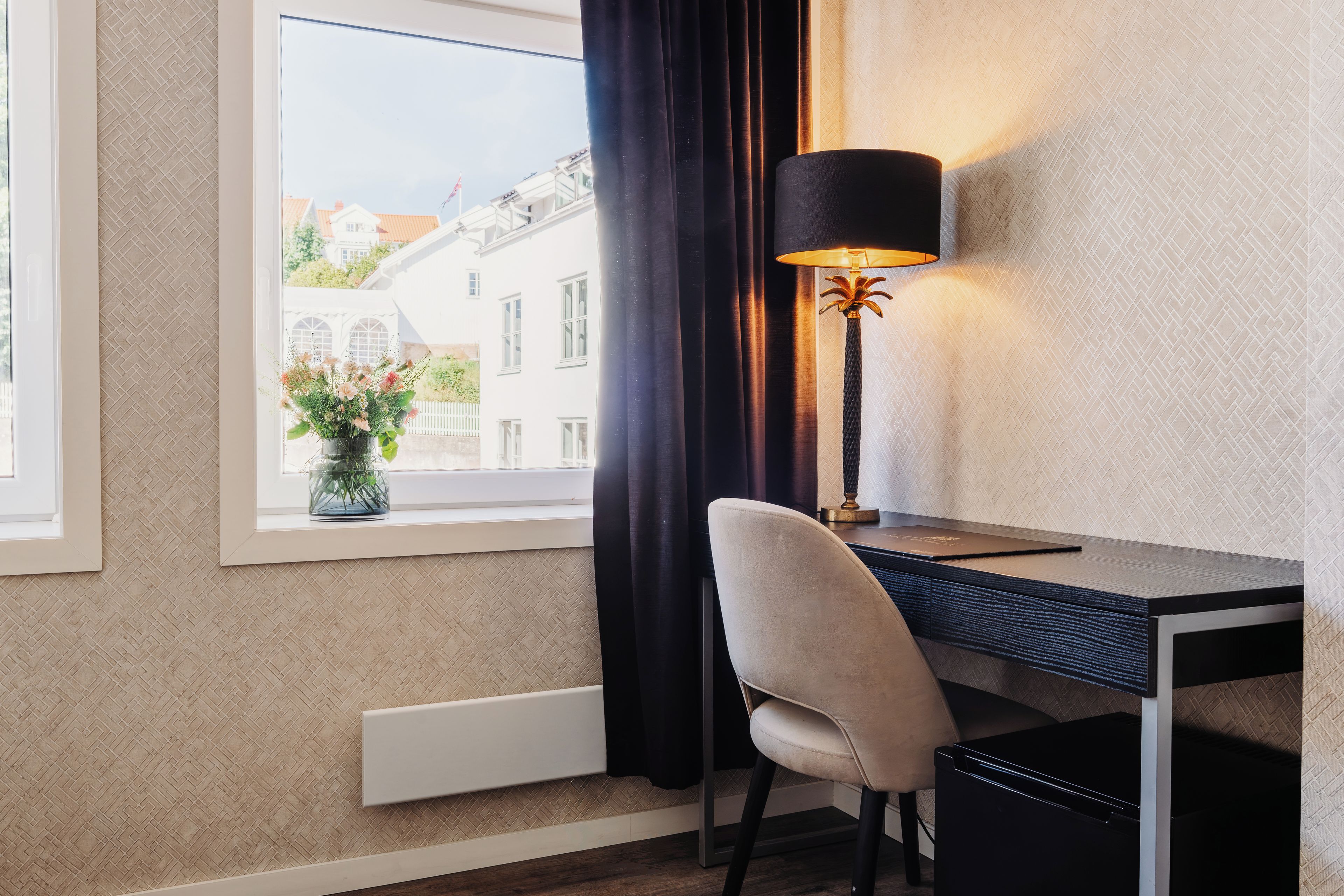 Habitación de hotel con escritorio, silla, lámpara y vistas a edificios a través de una ventana.