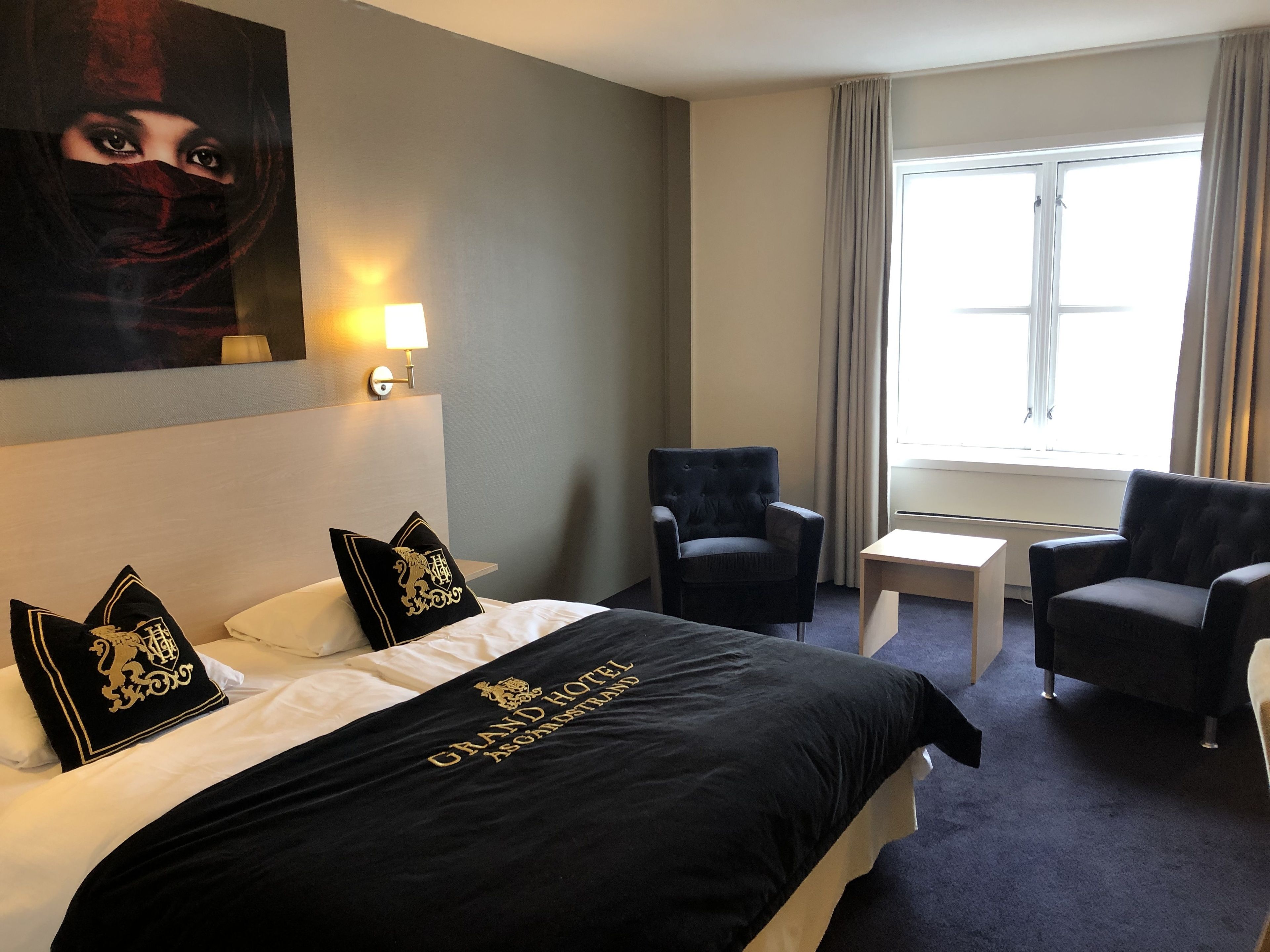 Una habitación de hotel con una cama, dos sillones, una mesita, una lámpara y una ventana con cortinas.