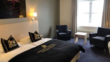 Una habitación de hotel con una cama, dos sillones, una mesita, una lámpara y una ventana con cortinas.