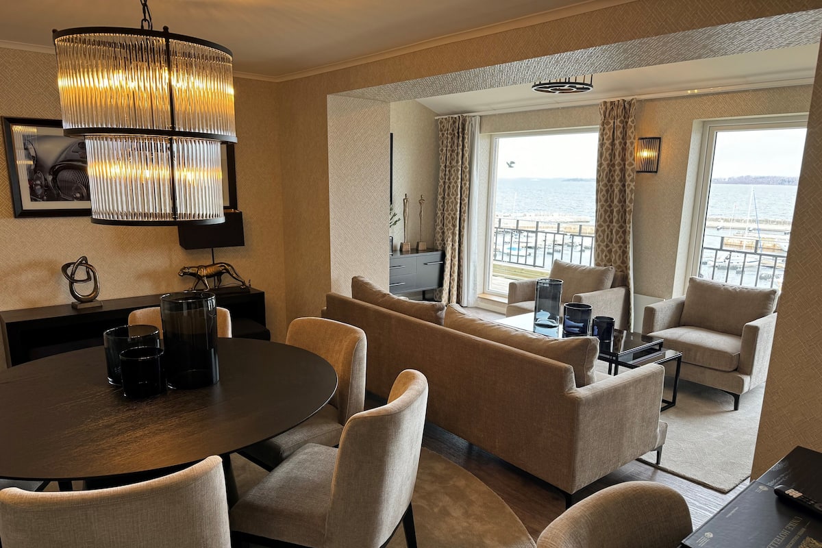 A modern hotel room with a round dining table, beige sofas, a large window with a view of the sea, and a chandelier.