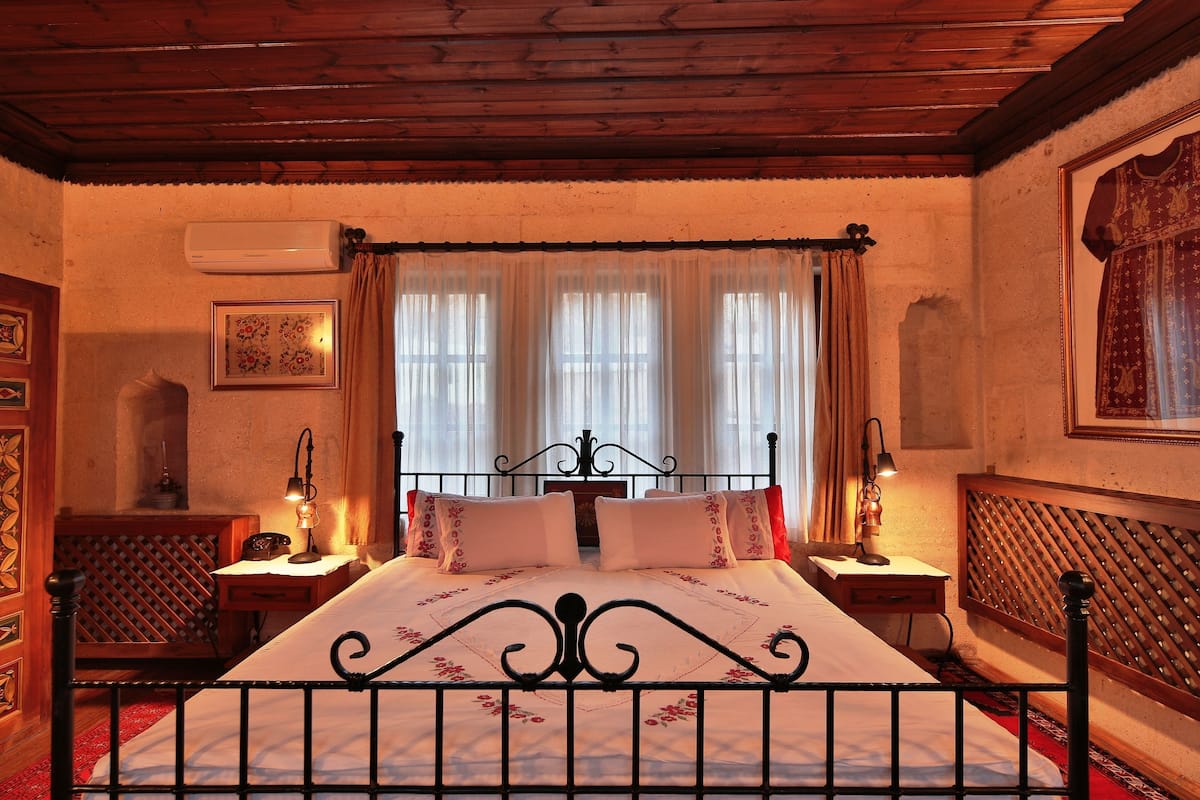 Un dormitorio tradicional con techo de madera, cama con estructura metálica, dos mesitas de noche con lámparas y un ventanal con cortinas.