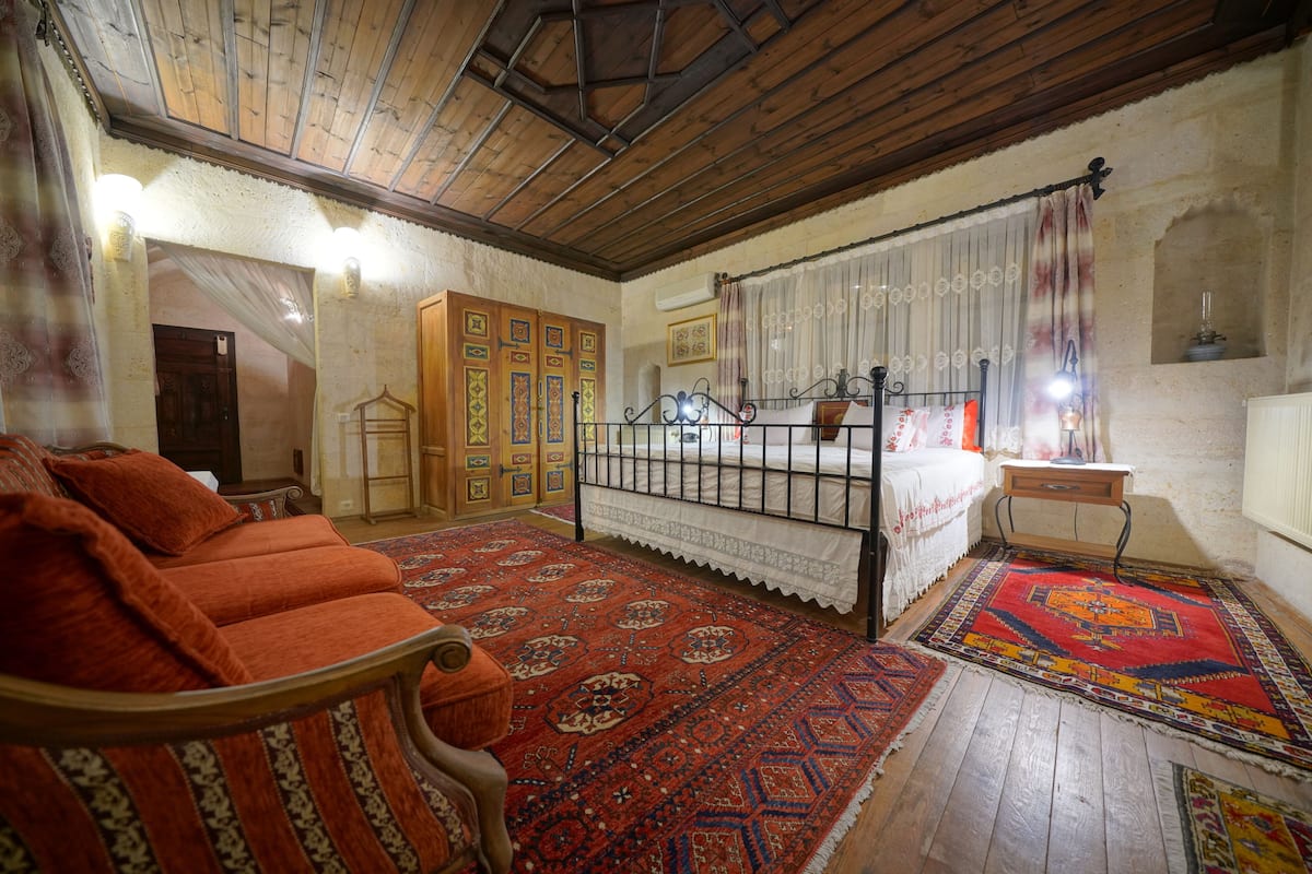 Una habitación tradicional con techo de madera, una cama con colcha estampada rojo y blanco, un sofá rojo y una alfombra grande con diseño.