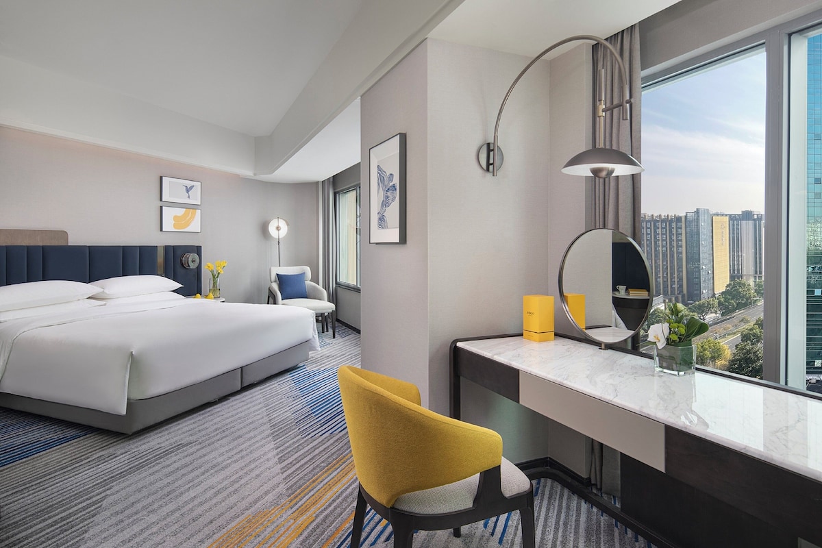 Quarto de hotel com uma cama, escrivaninha com espelho, uma cadeira e vista para a paisagem urbana.
