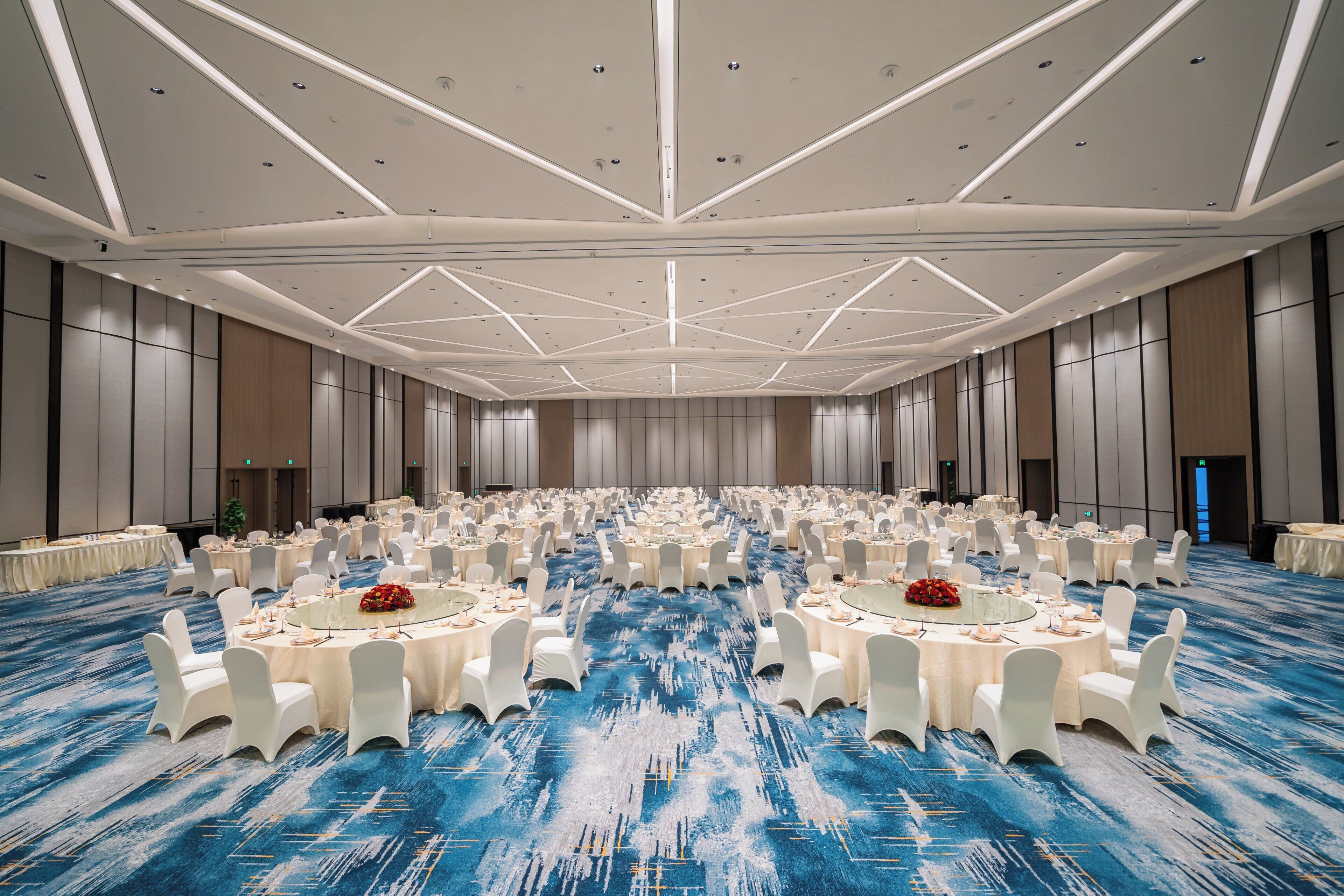 Een ruime balzaal met ronde tafels en witte stoelen, een blauw tapijt en een hoog plafond met ingebouwde verlichting.