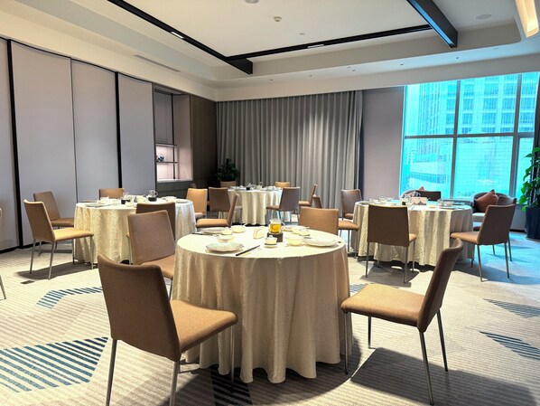 A hotel conference room with round tables, chairs, and a large window with a city view.