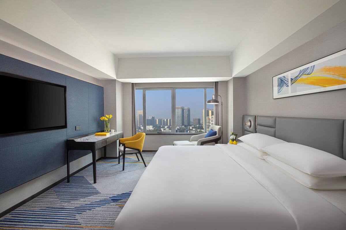 Habitación de hotel con cama, escritorio, silla, televisión y un ventanal con vistas a la ciudad.