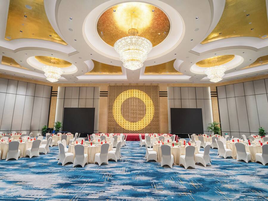 Un amplio salón de conferencias con mesas redondas, sillas blancas y un escenario con una pantalla grande.