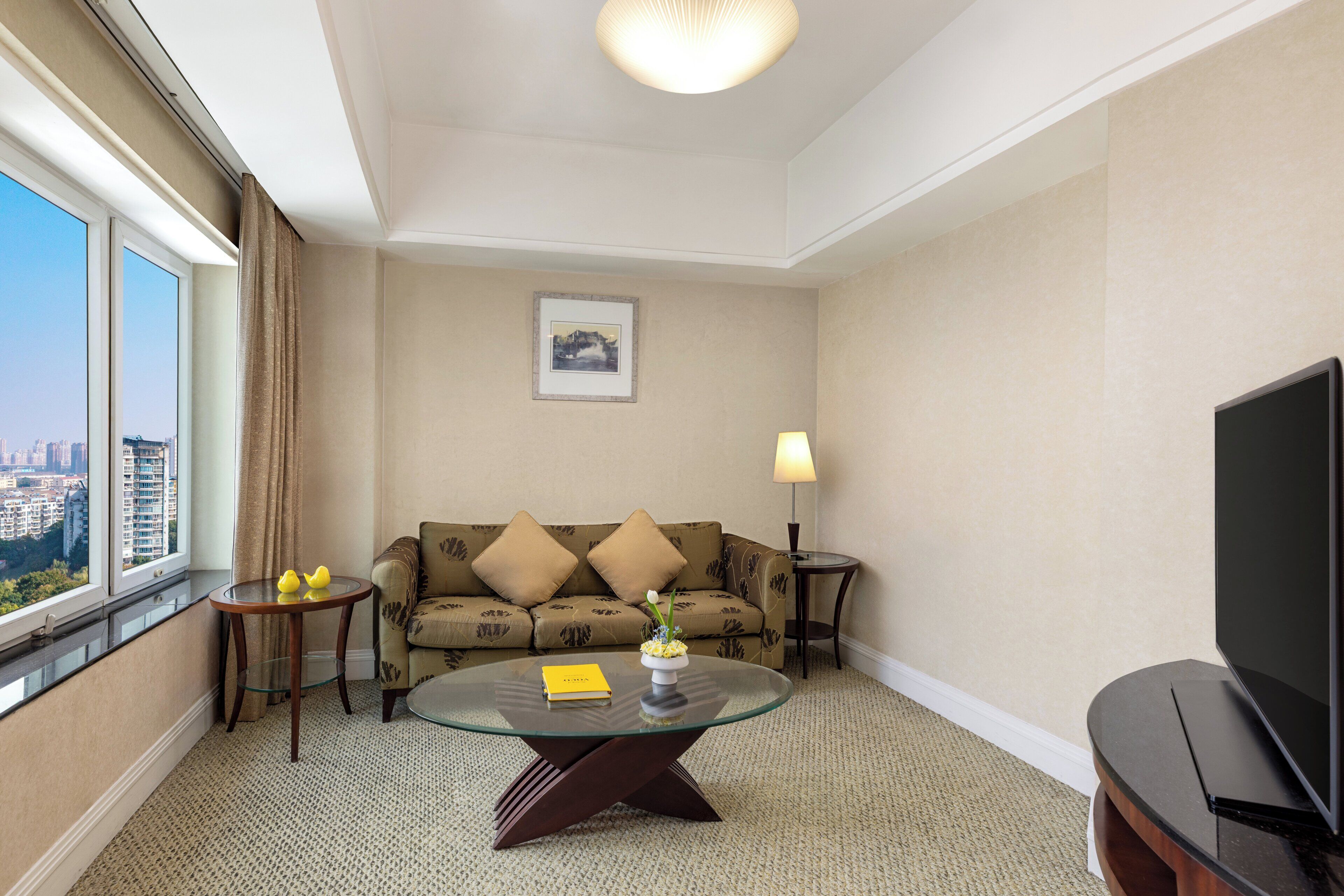 A hotel room with a sofa, a glass coffee table, a lamp, a TV, and a view of the city through the window.