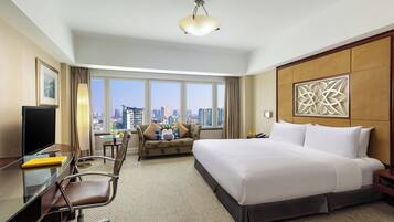 A hotel room with a large bed, a desk with a TV, a sofa, and a view of the cityscape.