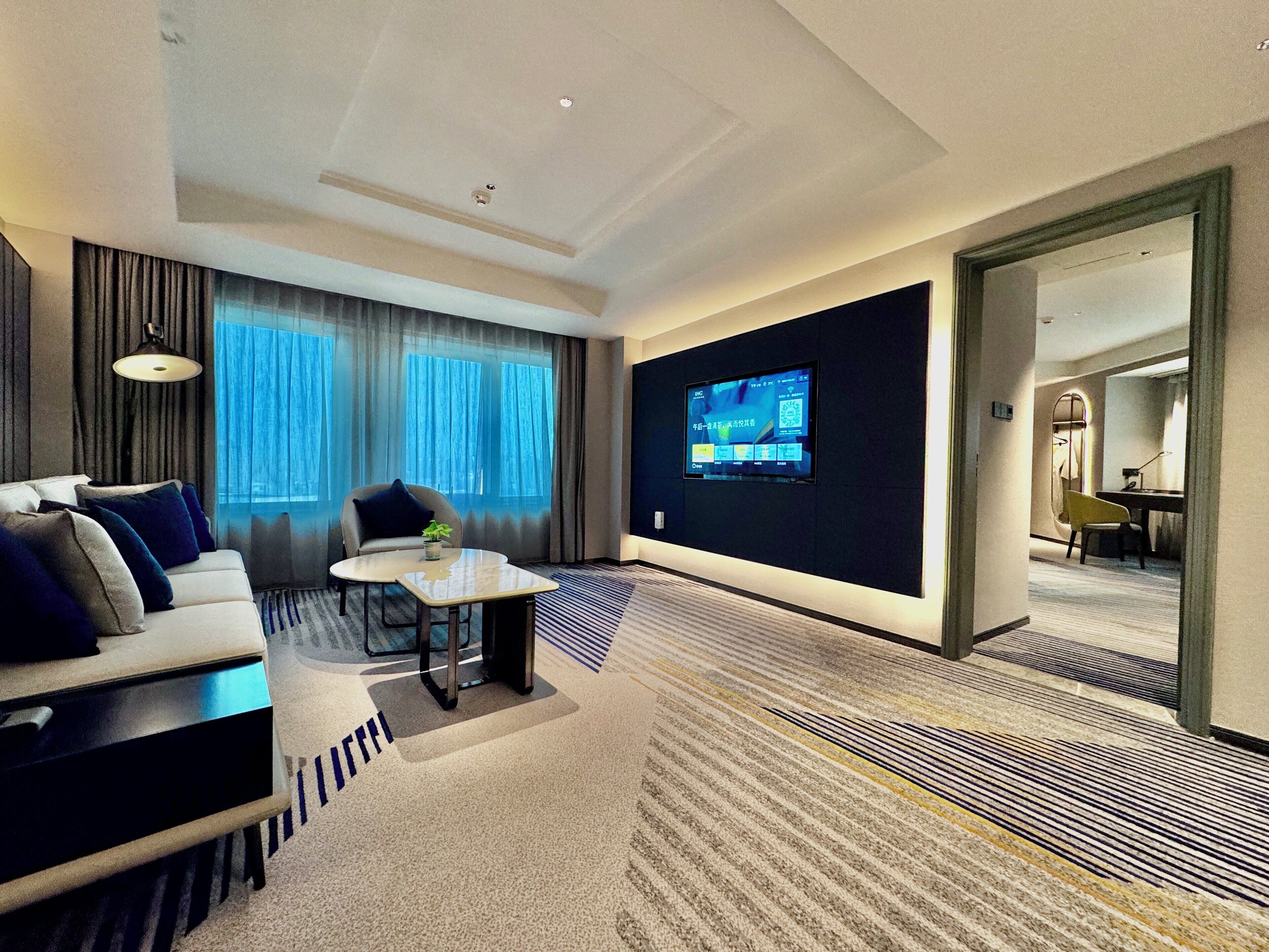 Quarto de hotel com sofá, mesa pequena, TV fixada na parede e vista para outro quarto por uma porta aberta.