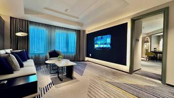 Quarto de hotel com sofá, mesa pequena, TV fixada na parede e vista para outro quarto por uma porta aberta.