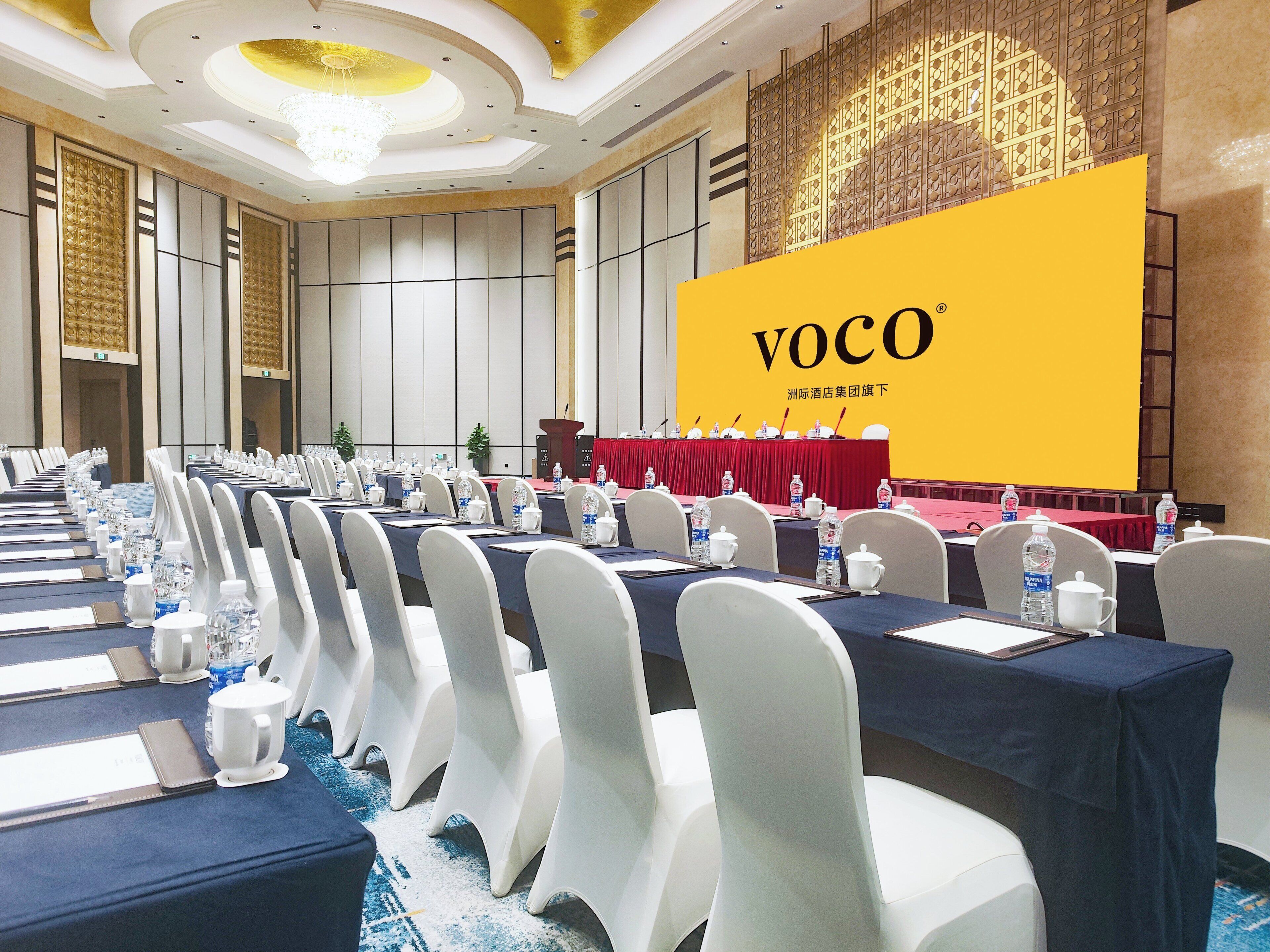 Un salón de conferencias con filas de mesas, sillas y una pantalla grande que muestra 'VOCO'.