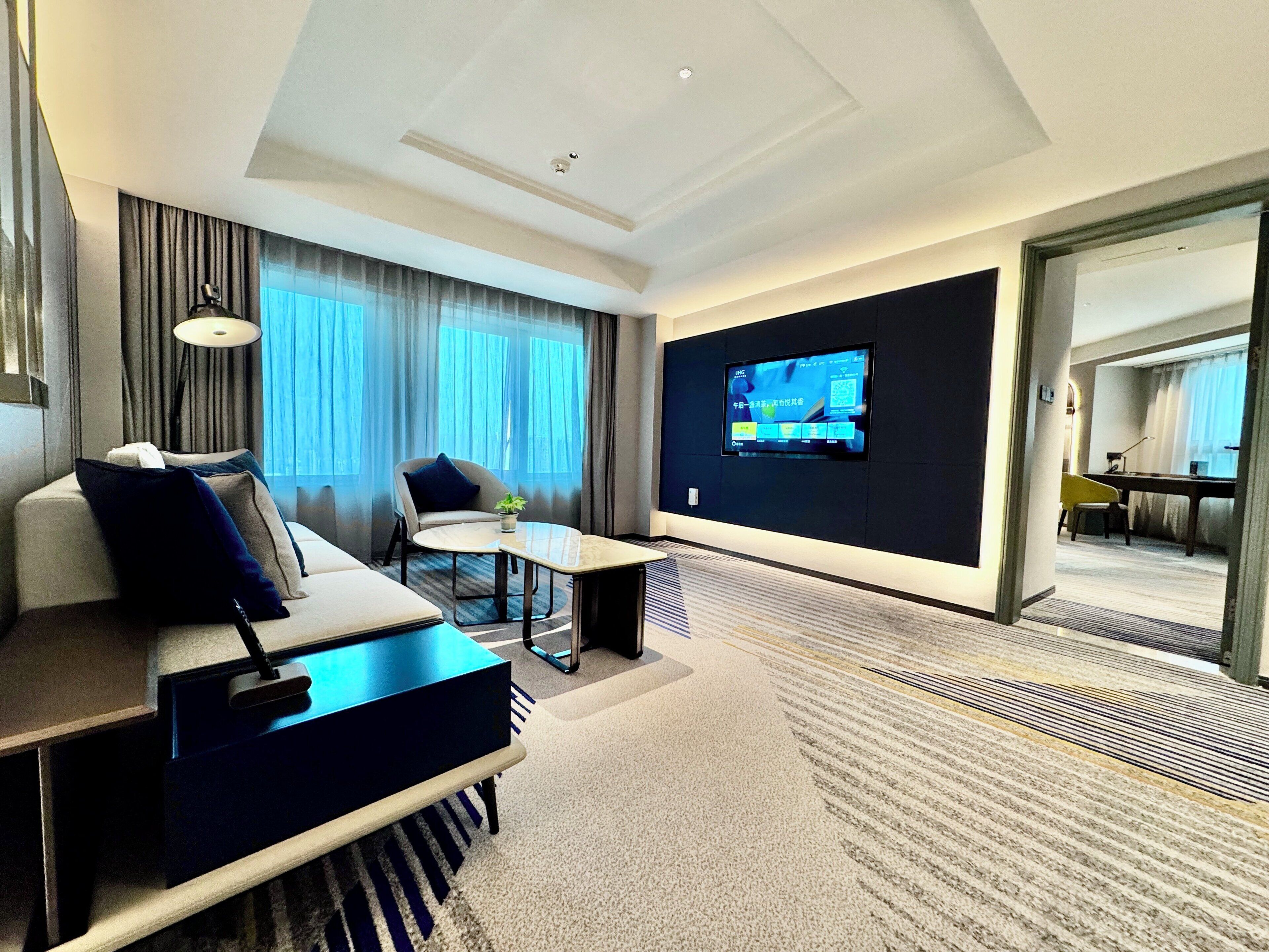 A modern hotel room with a sofa, armchair, small table, TV, and a view through an open door.
