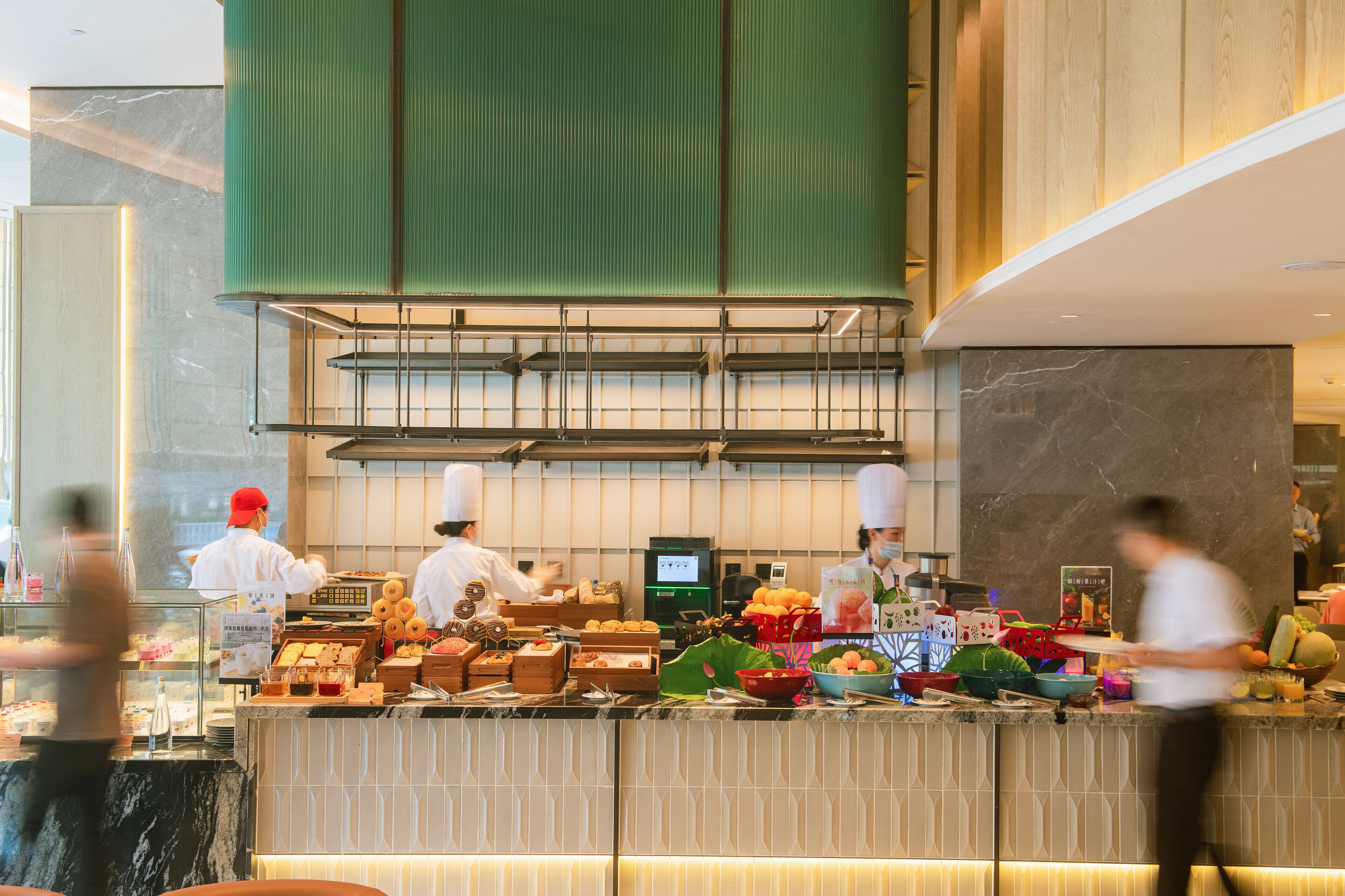 Una cocina de hotel con cocineros preparando alimentos, una zona de bufé con diversos platos y un diseño de interiores moderno.