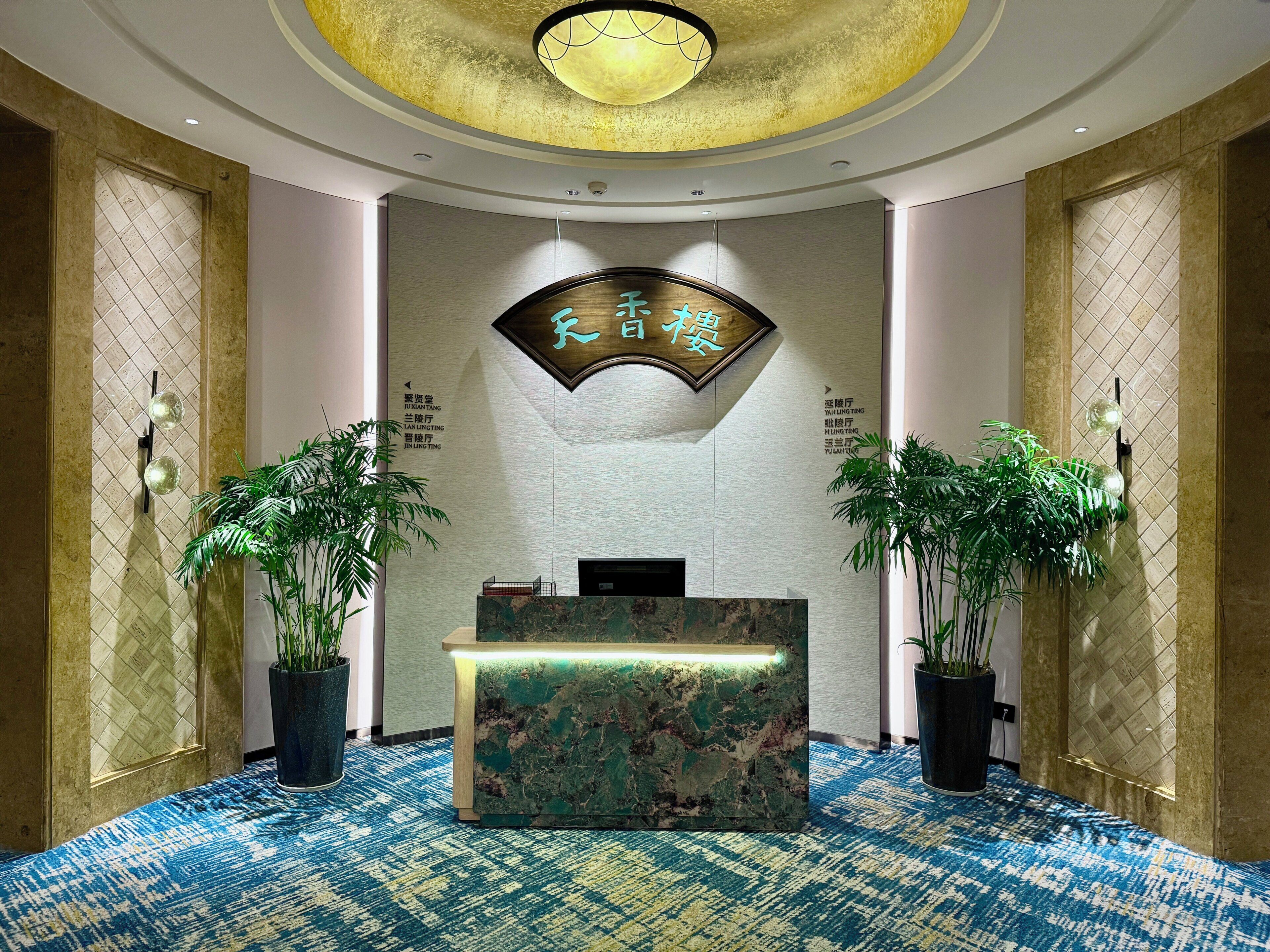 A reception desk with a marble top, flanked by two potted plants, in a lobby with patterned carpet and a decorative ceiling.