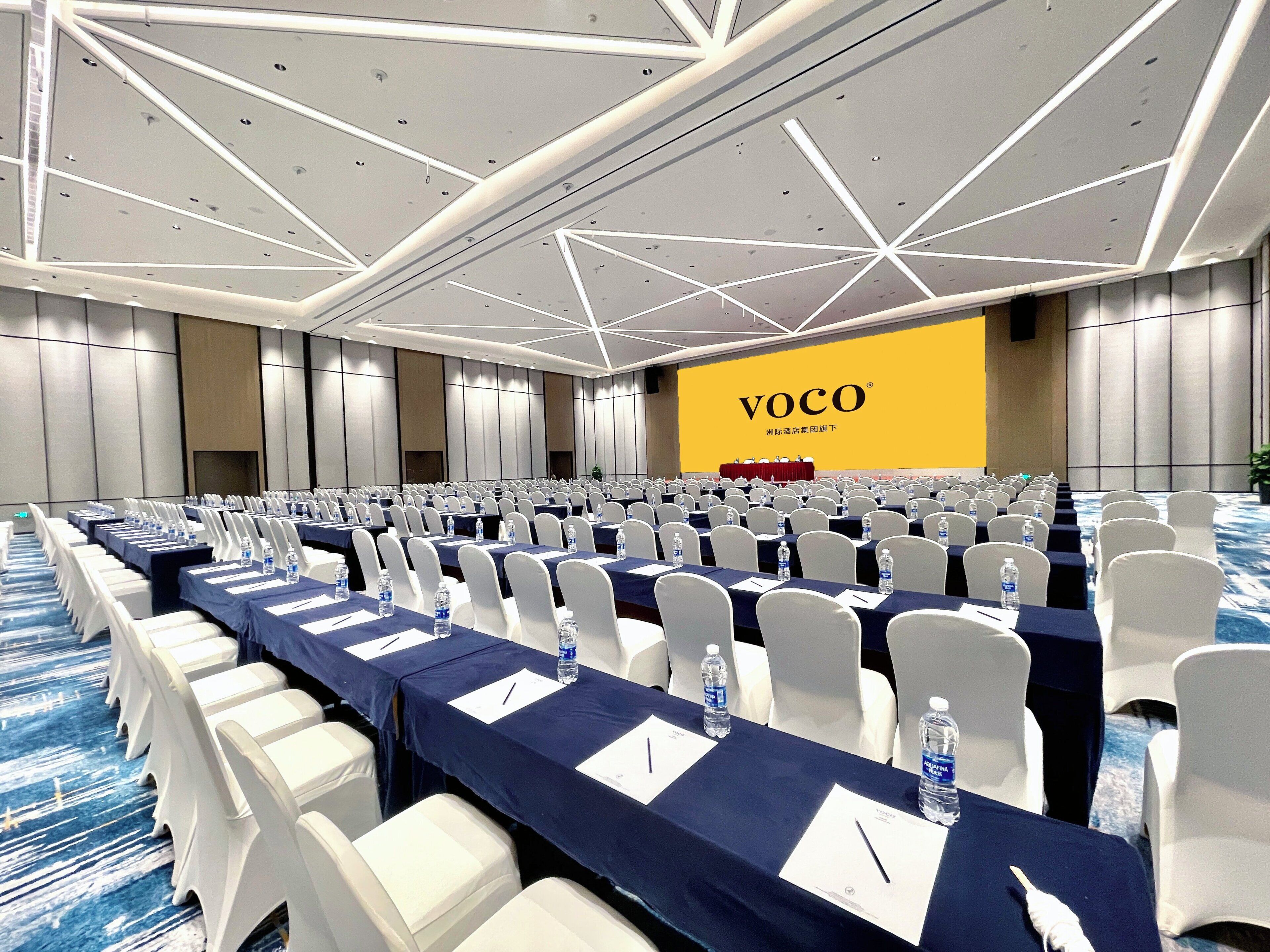 Una sala conferenze con file di tavoli, sedie e un grande schermo che mostra 'VOCO'.