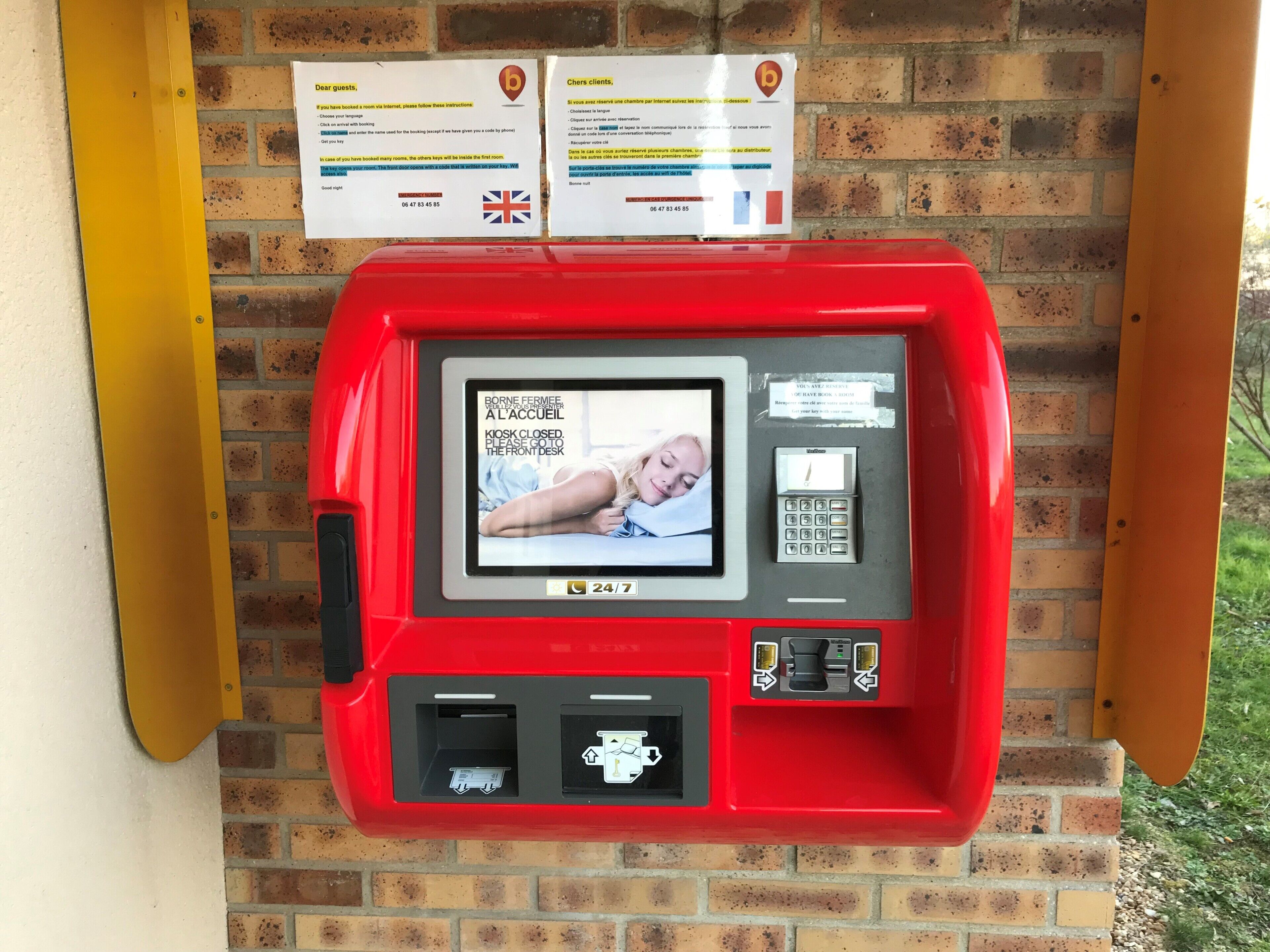 Un kiosque rouge en libre-service, doté d’un écran affichant un message indiquant que le comptoir d’accueil est fermé, ainsi que des instructions à l’intention des clients.