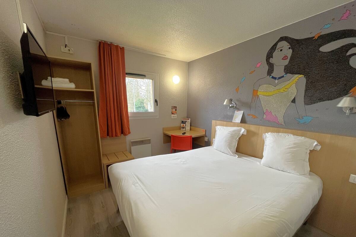 Ein Hotelzimmer mit einem großen Bett, einem Schreibtisch mit Stuhl, einem kleinen Tisch, einem Regal mit Handtüchern, einem Fenster mit Vorhängen und einem Wandgemälde.