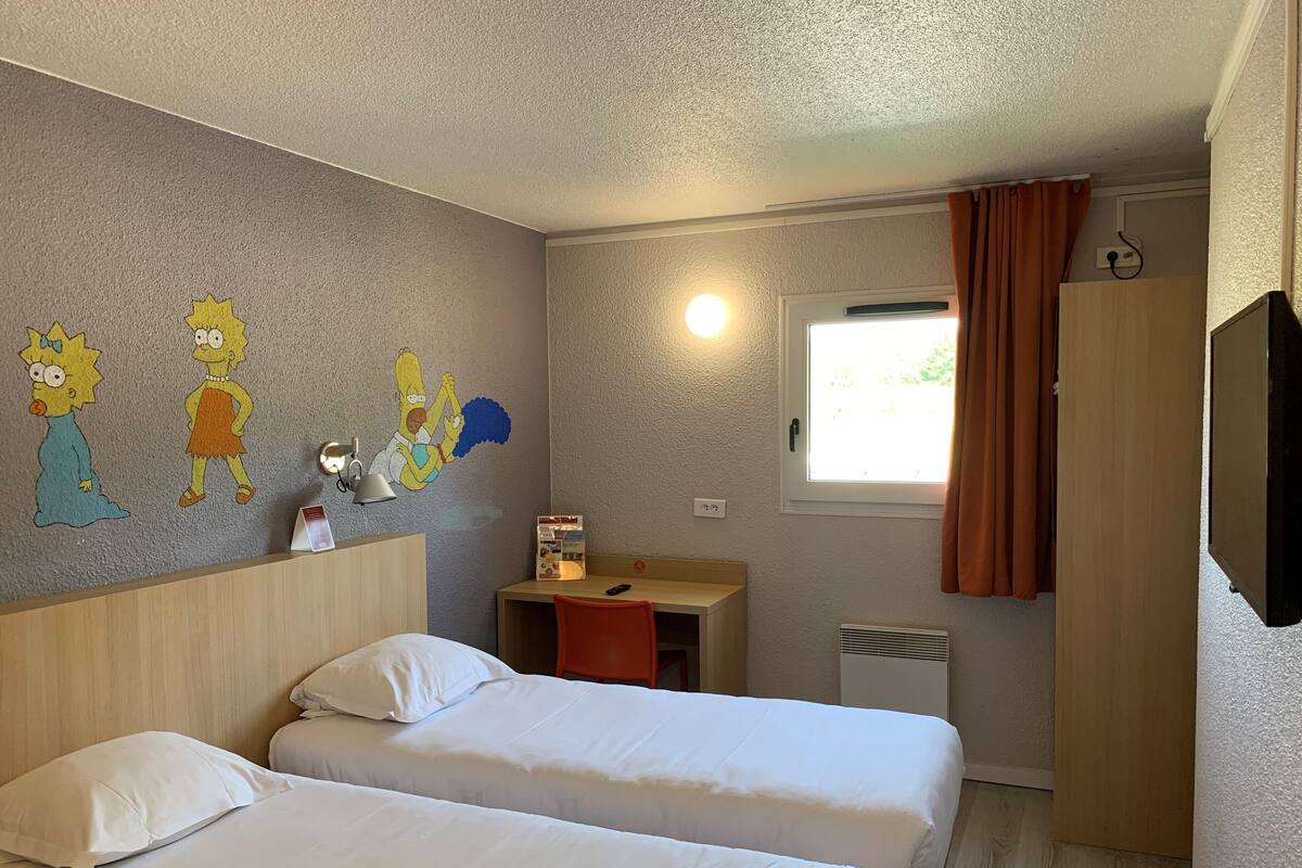Ein Hotelzimmer mit zwei Betten, einem Wandgemälde, einem Schreibtisch mit Lampe, einem Fenster mit Vorhängen und einem Fernseher an der Wand.