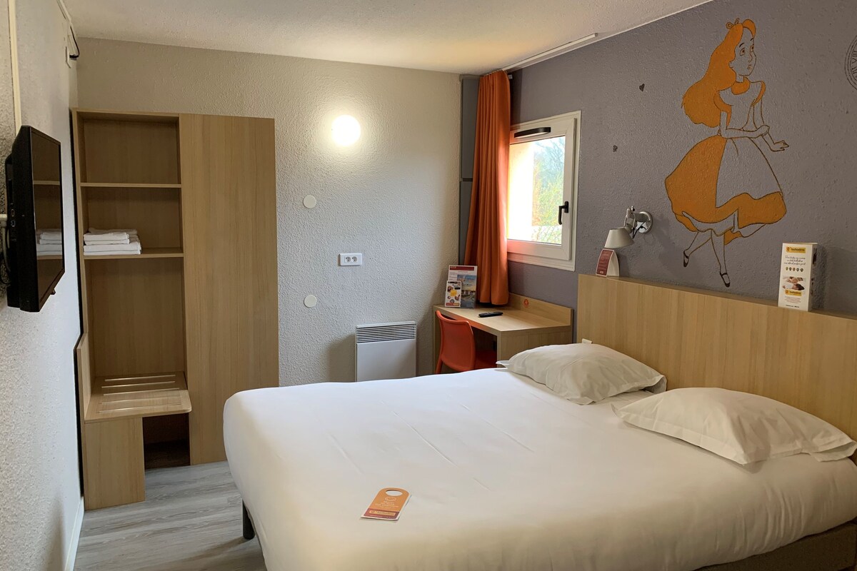 Ein Hotelzimmer mit Bett, Schreibtisch, Kleiderschrank und einem an der Wand befestigten Fernseher.