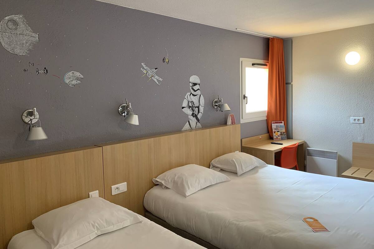 Ein Hotelzimmer mit zwei Betten, einer Wand mit Star Wars-Dekorationen, einem Nachttisch mit Lampe und einem Fenster mit orangenen Vorhängen.