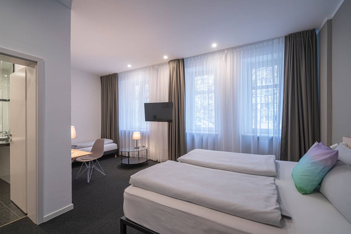 A hotel room with two beds, a desk, a chair, a TV, and large windows with curtains.