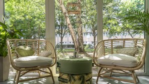 Two wicker chairs with cushions, a small round table with a decorative item, and a view of trees and buildings through large windows. -  BIG MAMA Leipzig (Leipzig)