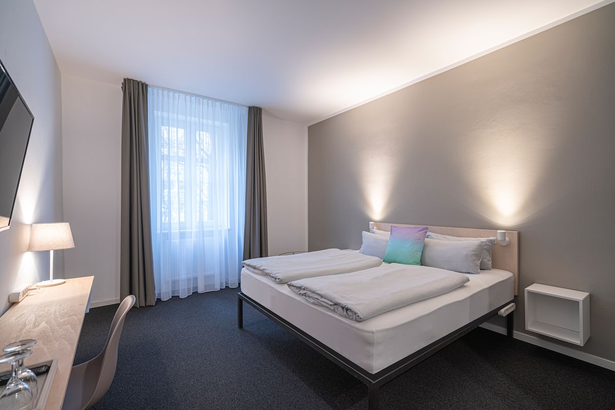 A hotel room with a bed, bedside tables, a desk with a lamp, and a TV.