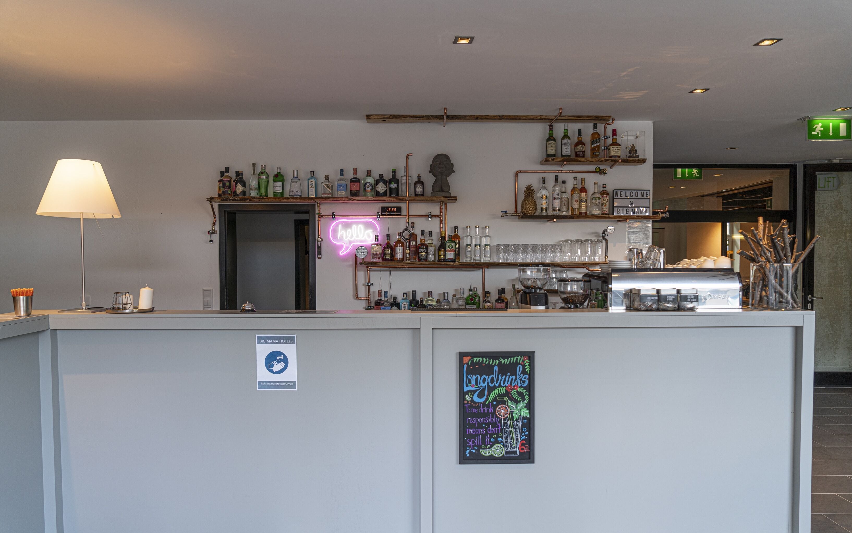 Un espace bar avec des étagères remplies de bouteilles d’alcool, un panneau néon et un tableau noir sur lequel est écrit un message.