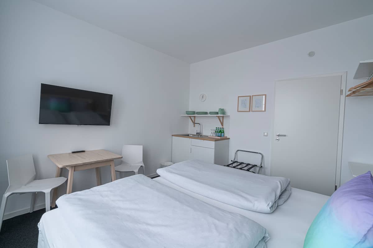 A hotel room with two beds, a desk, a chair, a TV, a small kitchenette, and a door.