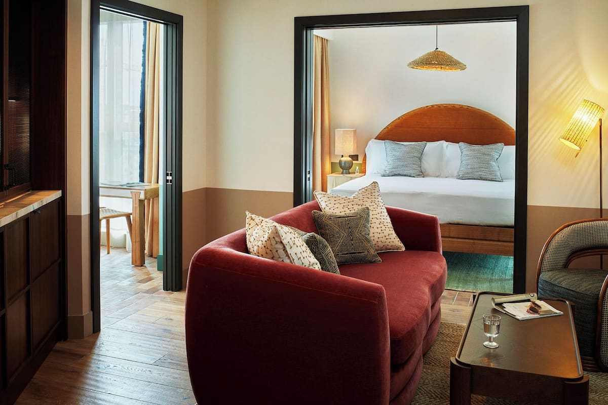 A hotel room with a bed, a red armchair, a small table, and a mirror reflecting the room.