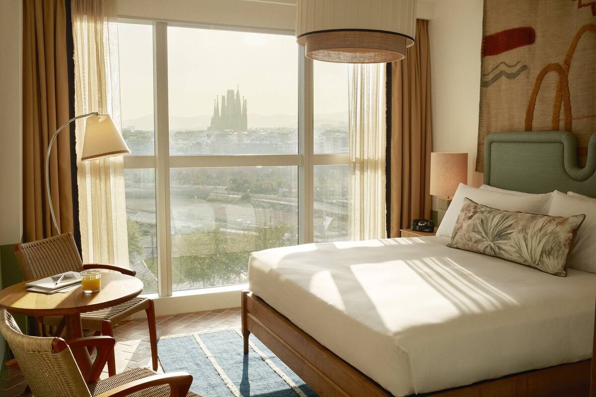 A hotel room with a large window, a bed, a chair, a table, and a lamp.