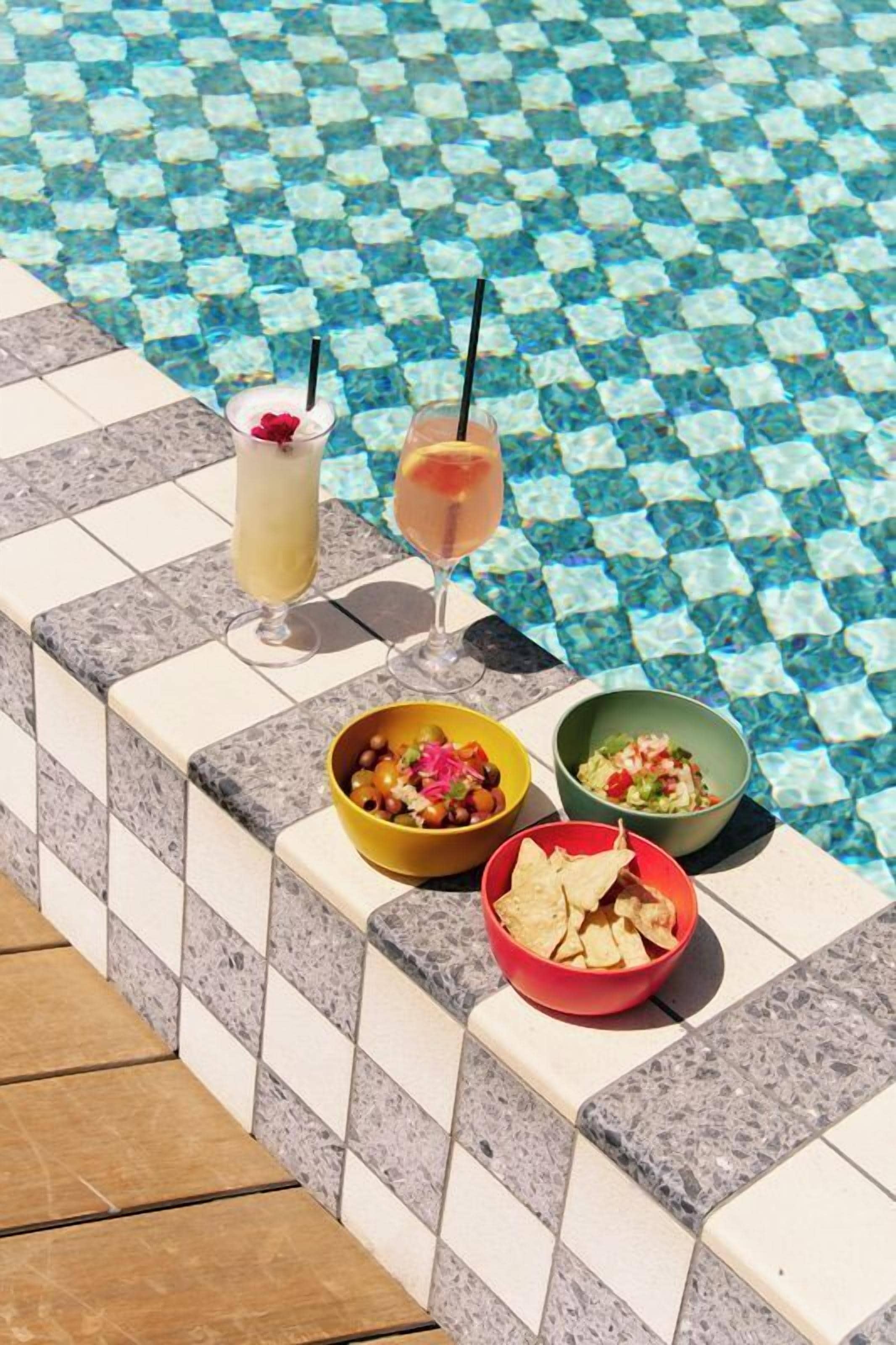 A poolside setting with a tiled edge, two drinks, and bowls of food.