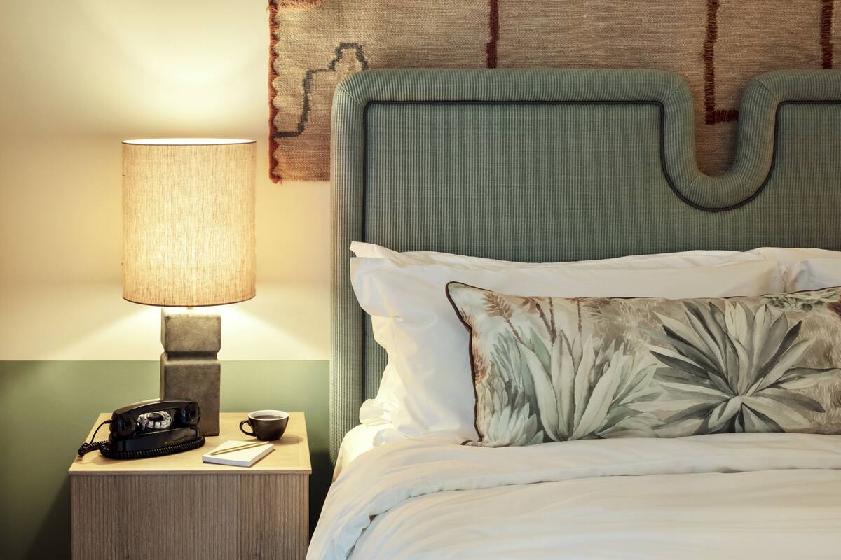 A neatly made bed with a floral pillow, a bedside lamp, a vintage telephone, and a cup of coffee on a wooden nightstand.