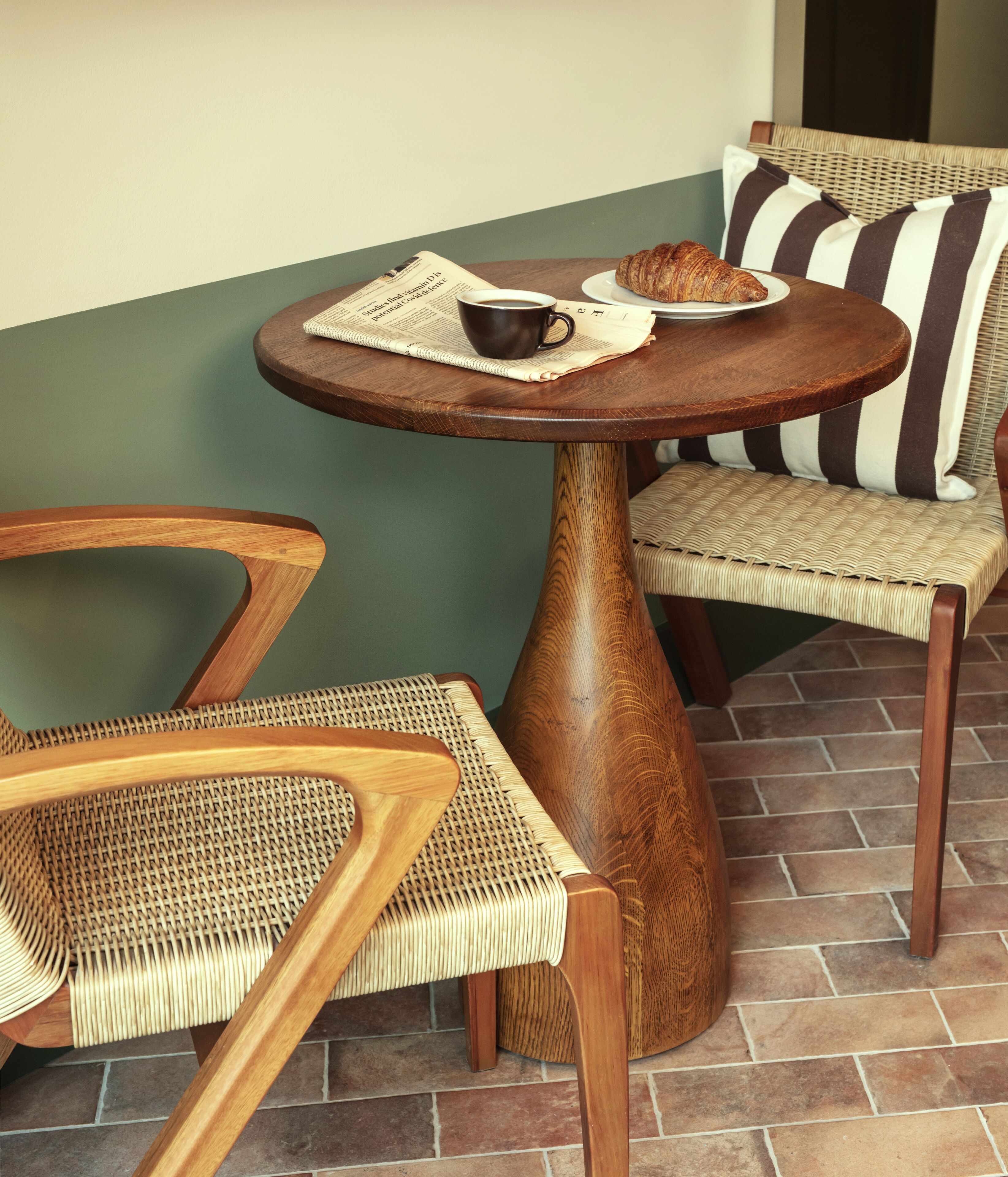 Una pequeña mesa redonda de madera con un periódico, una taza de café y un croissant sobre una silla de mimbre.